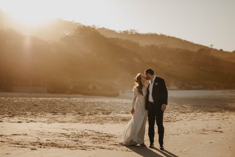 Bride and groom walking on beach at Broken Head with the sun shine on them as they kiss