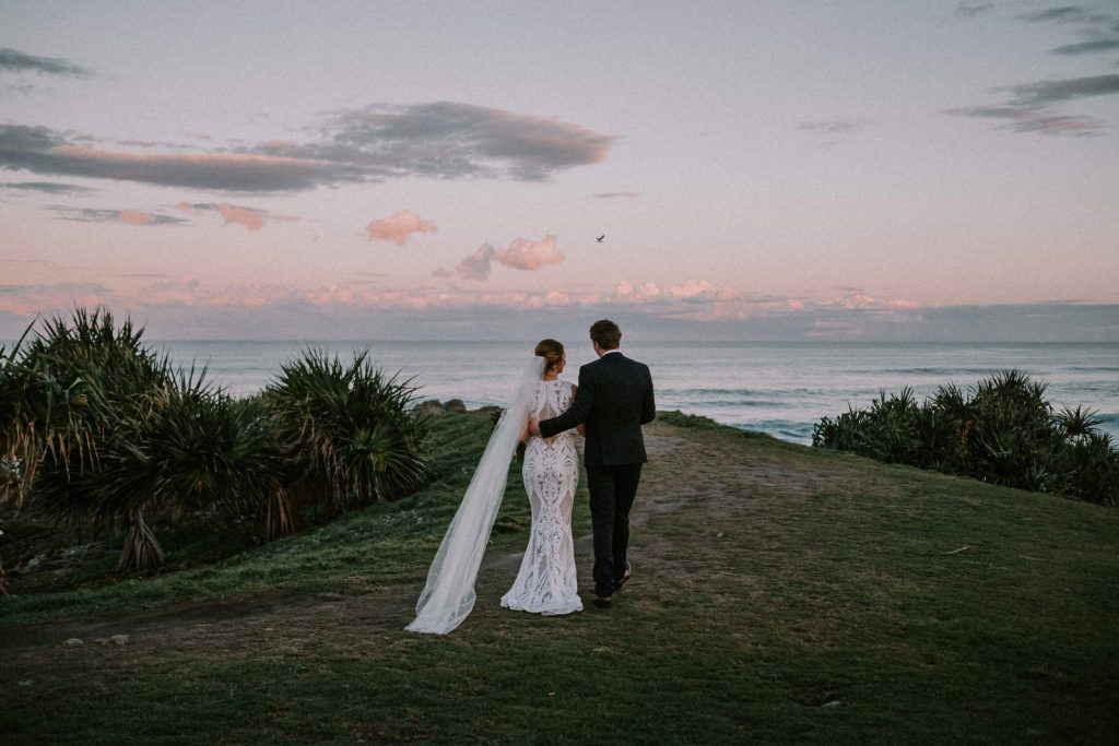 Gold Coast Wedding photographer, bride and groom in wedding attire on their wedding day. Walking towards the ocean on a point in each other's arms. The sun sets with pink hues in the sky