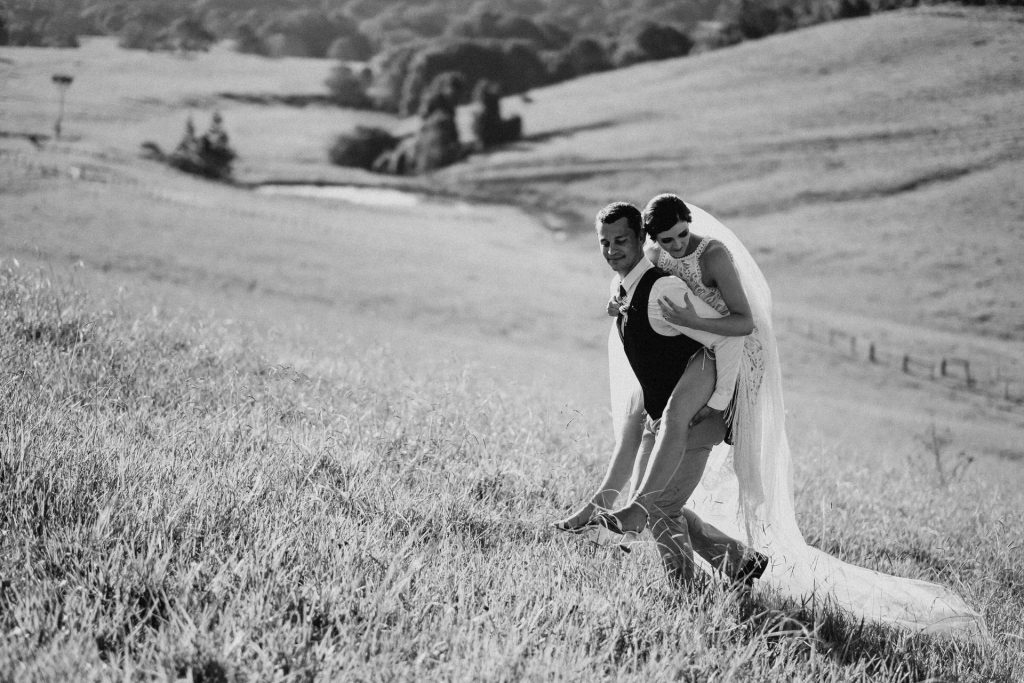 Byron Bay wedding photographer, Groom giving a bride a piggy back up the hill on their wedding day. The brides veil flows down trailing behind both of them as she holds the groom tight.