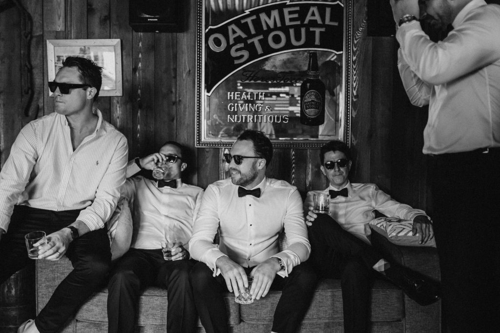 Byron Bay Wedding, Groomsmen dressed in tuxedo shirts and black bow ties, sitting casually on a couch drinking whisky.