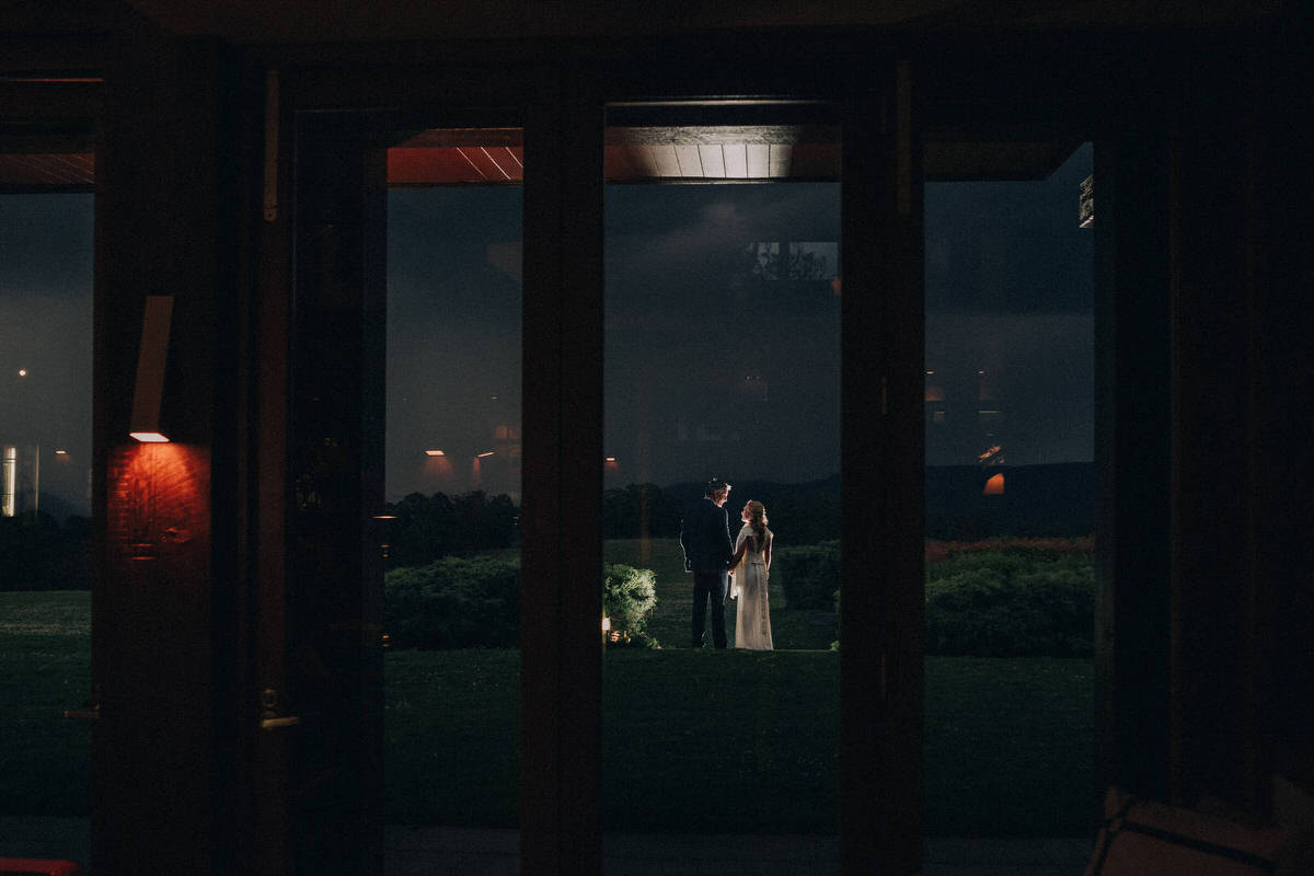 Looking through the large windows of spicers peak lodge at night as bride and groom stand outside looking at the stormy sky